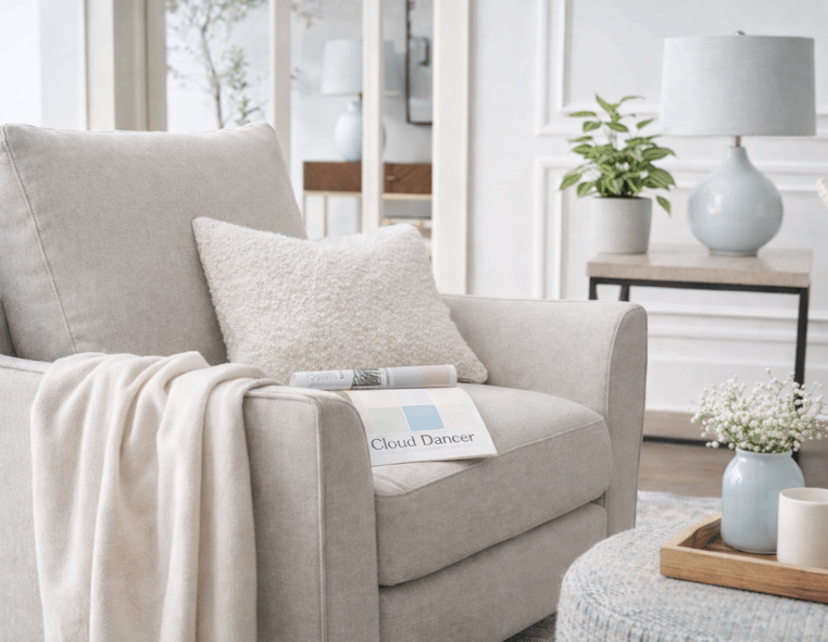Neutral accent chair styled with soft white textiles, a light wood side table, white lamp, and greenery inspired by Pantone’s 2026 Color of the Year, Cloud Dancer. | Furniture Mall