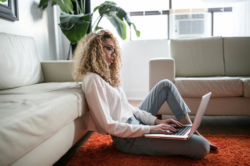Woman sitting on ground leaning against a couch surfing her laptop